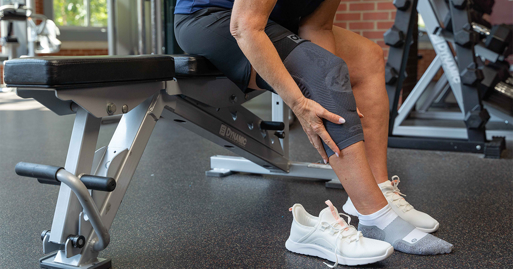 Woman sitting on workout bench while putting on her OrthoSleeve Compression Knee Brace and Socks